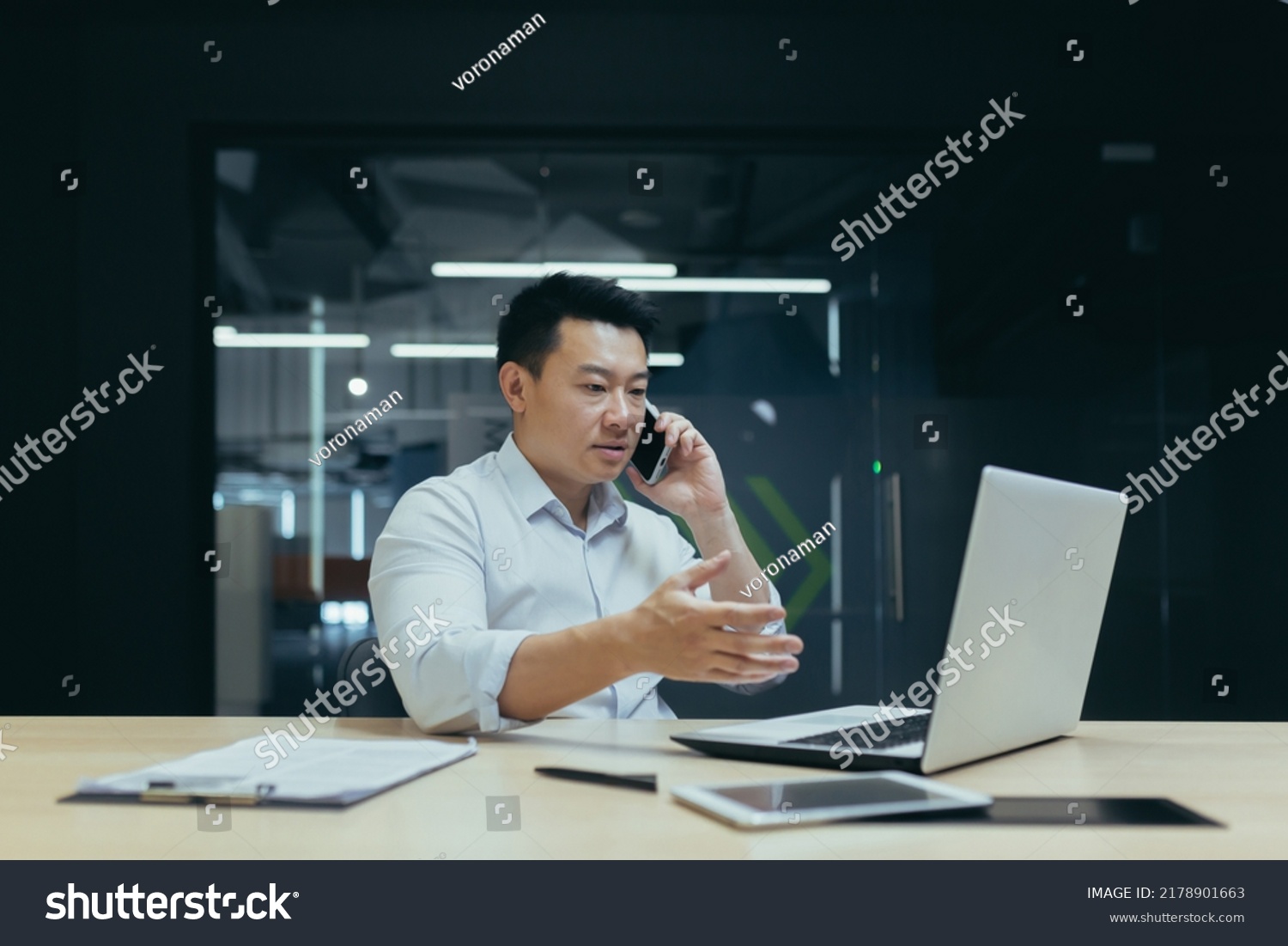 Worried and serious young male Asian businessman talking on the phone in the office. Sitting at a table with a laptop and documents. Trying to solve problems #2178901663