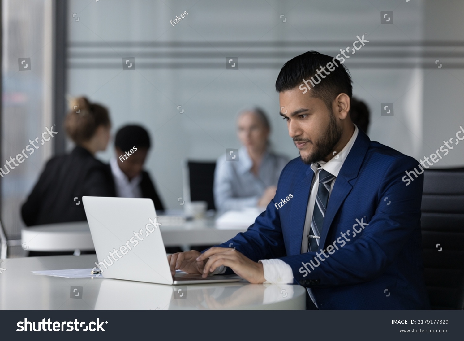 Focused Indian employee man working on online project at laptop, typing, chatting, sitting at workplace table in open office with business meeting of diverse colleagues in background #2179177829
