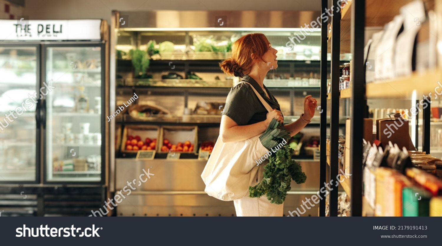 Female shopper choosing food products from a shelf while carrying a bag with vegetables in a grocery store. Young woman doing some grocery shopping in a trendy supermarket. #2179191413