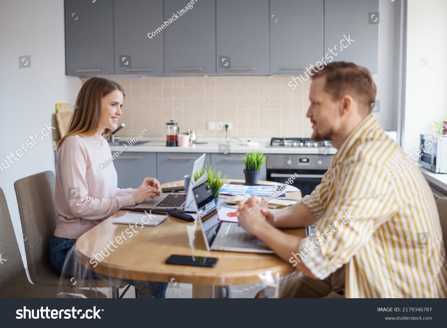 Side view of young couple working from home on laptops sitting together at table. Wife and husband discussing business while working remotely or as freelancers #2179346787