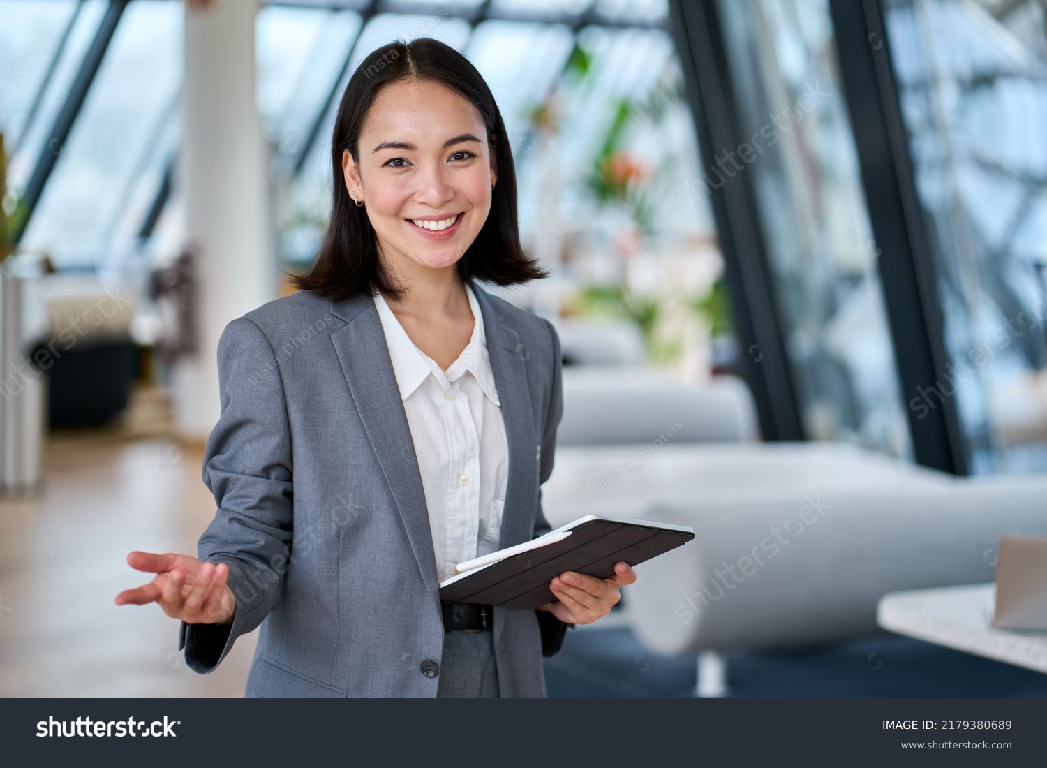Happy young Asian saleswoman looking at camera welcoming client. Smiling woman executive manager, secretary offering professional business services holding digital tablet standing in office. Portrait #2179380689