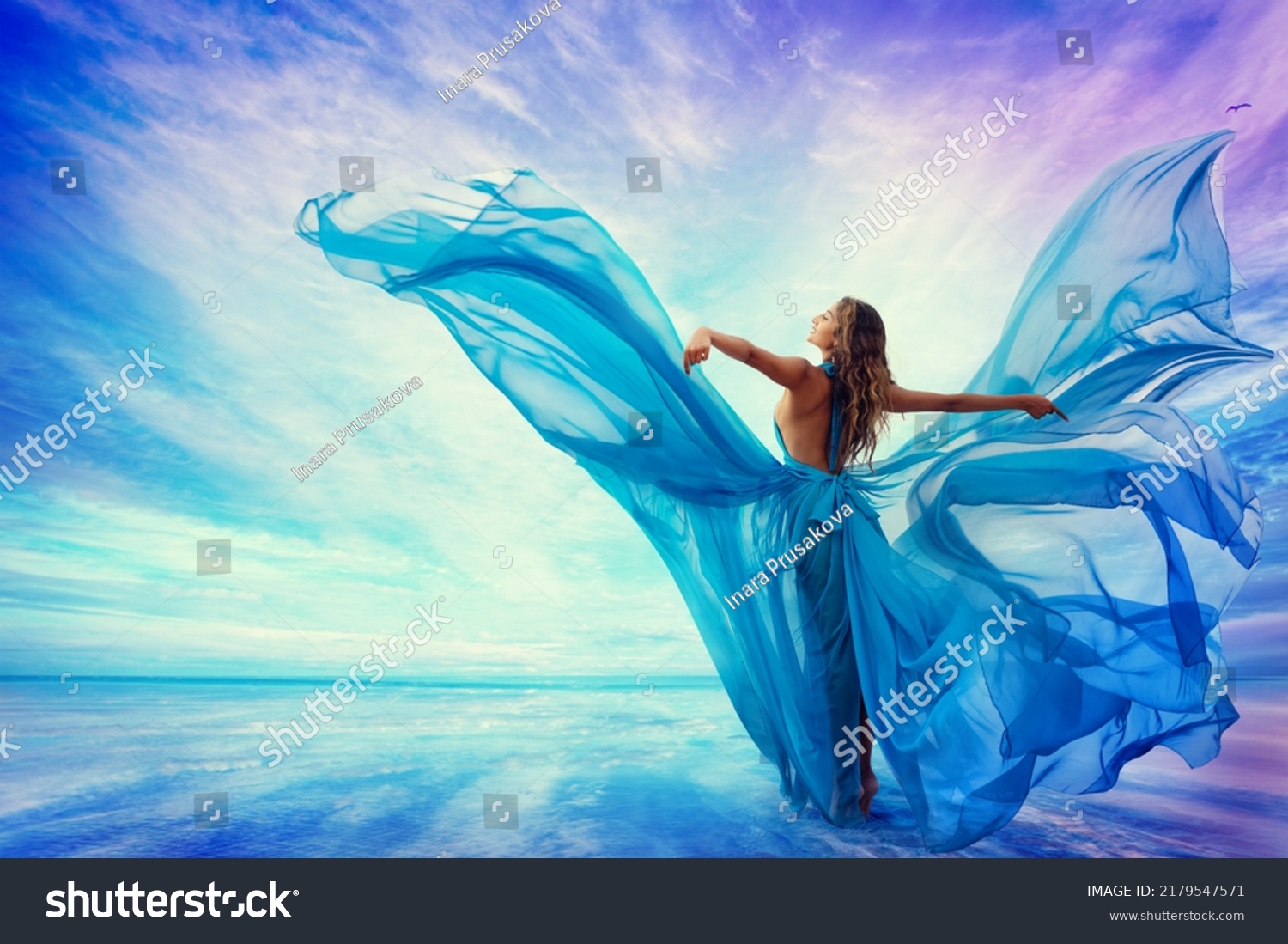 Woman in Blue Dress Flying on Wind looking at Sky and Sea. Beautiful Model Arms outstretched enjoying Freedom at Beach Summer Resort Rear view. Artistic Women Silhouette in Fantasy Gown as Butterfly #2179547571