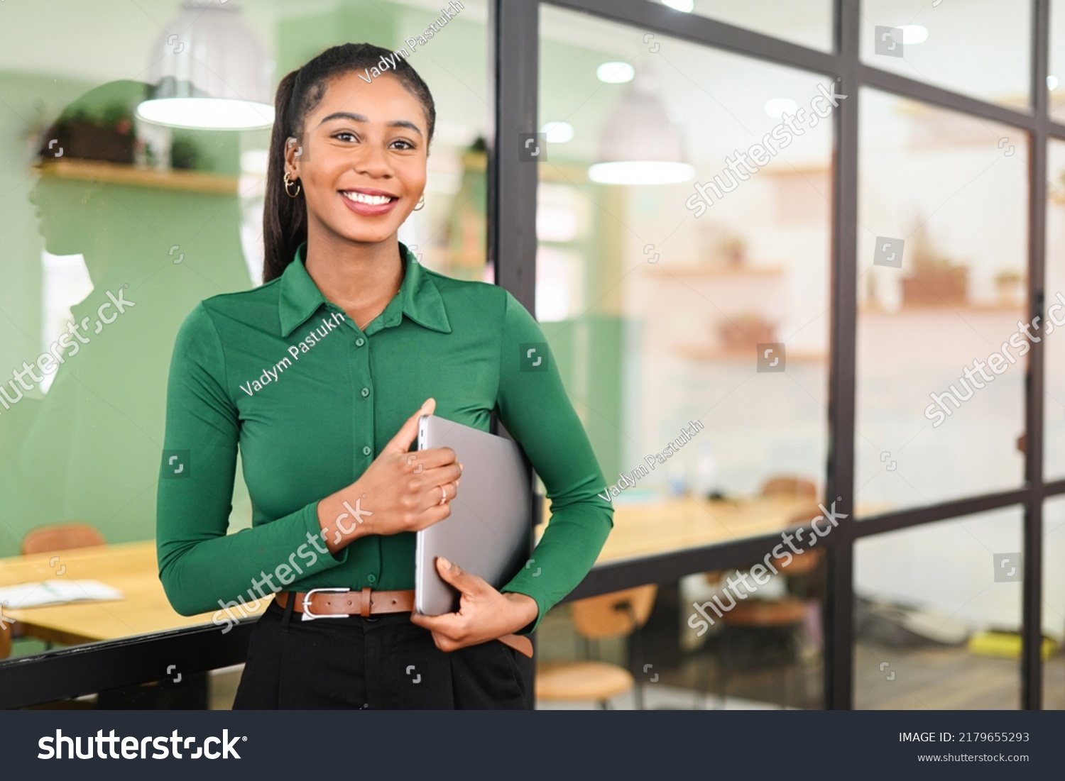 Carefree inspired african-american female employee standing in modern office space and holding laptop, cheerful black businesswoman in green shirt looks at the camera with light friendly smile #2179655293