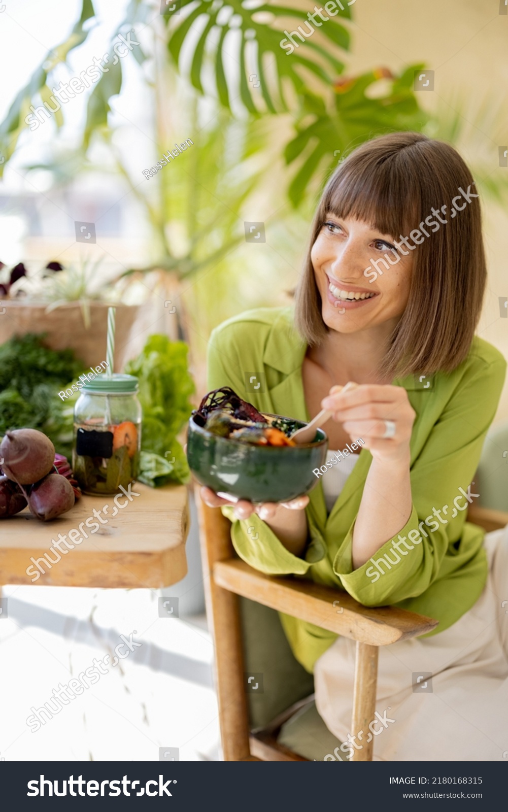 Young cheerful woman eats vegetarian lunch in bowl, sitting by the table full of fresh food ingredients indoors. Healthy lifestyle and wellness concept #2180168315