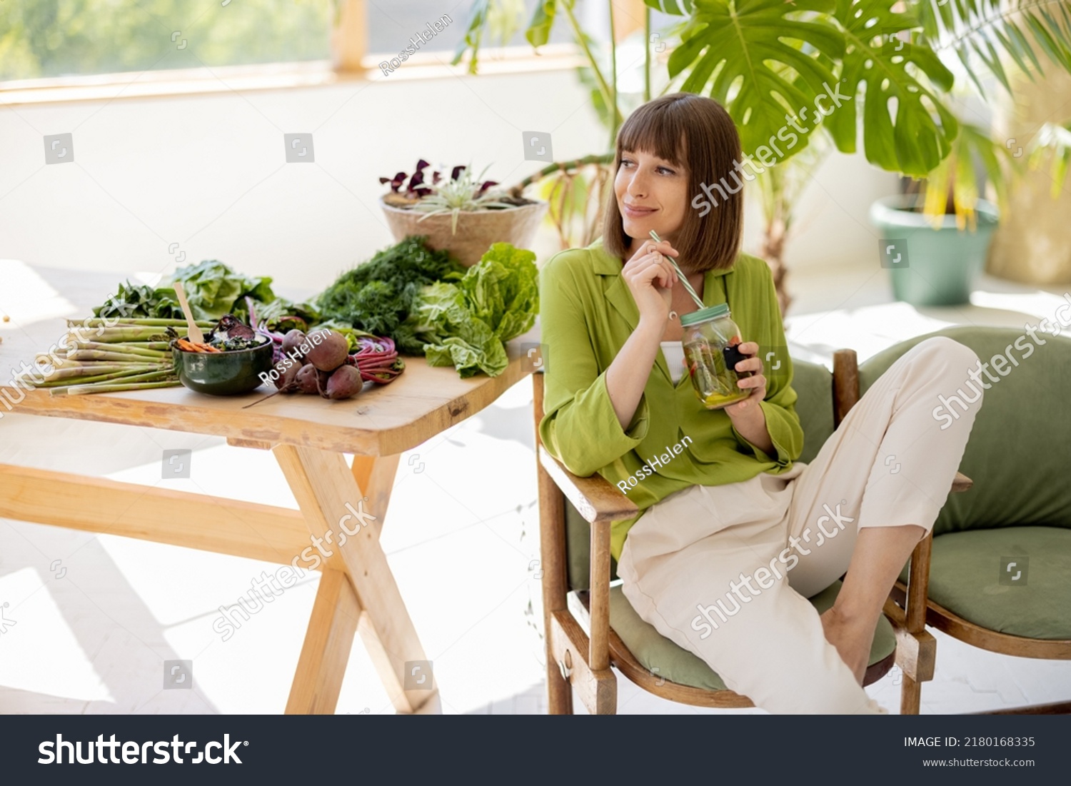 Young woman drinks lemonade while sitting on chair near table with lots of fresh food ingredients in room with green plants. Healthy lifestyle concept #2180168335