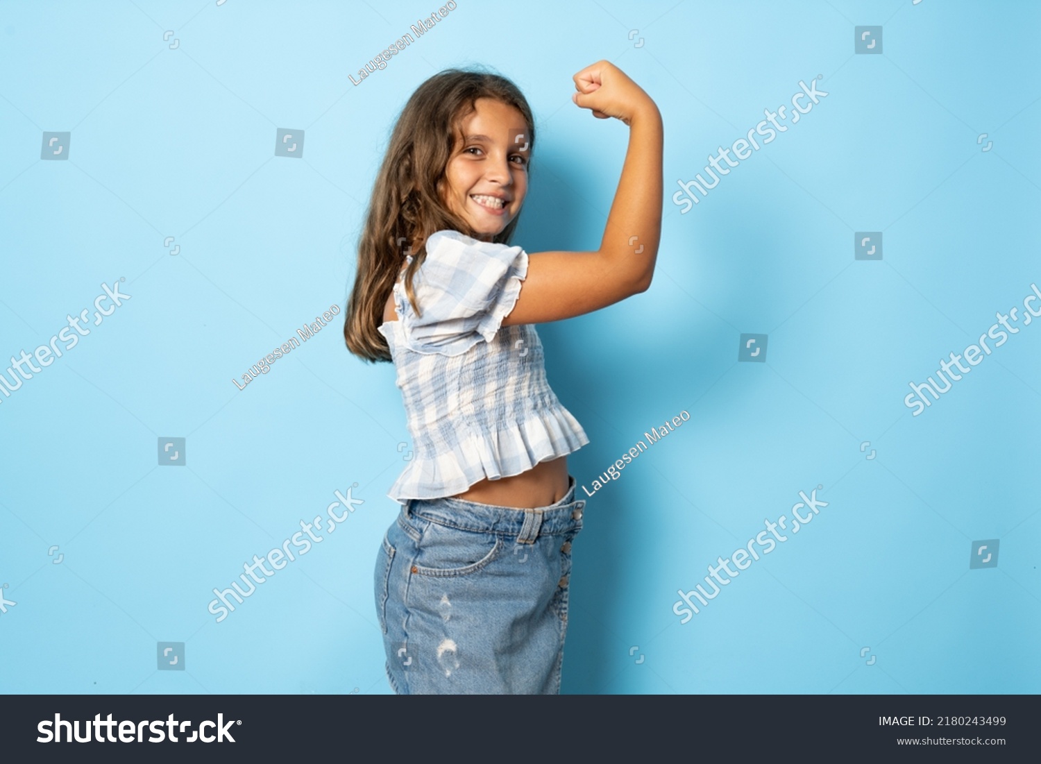 Smiling strong little brunette kid girl 10-11 years old showing biceps, muscles isolated on blue background children studio portrait. Childhood lifestyle concept. Mock up copy space. #2180243499
