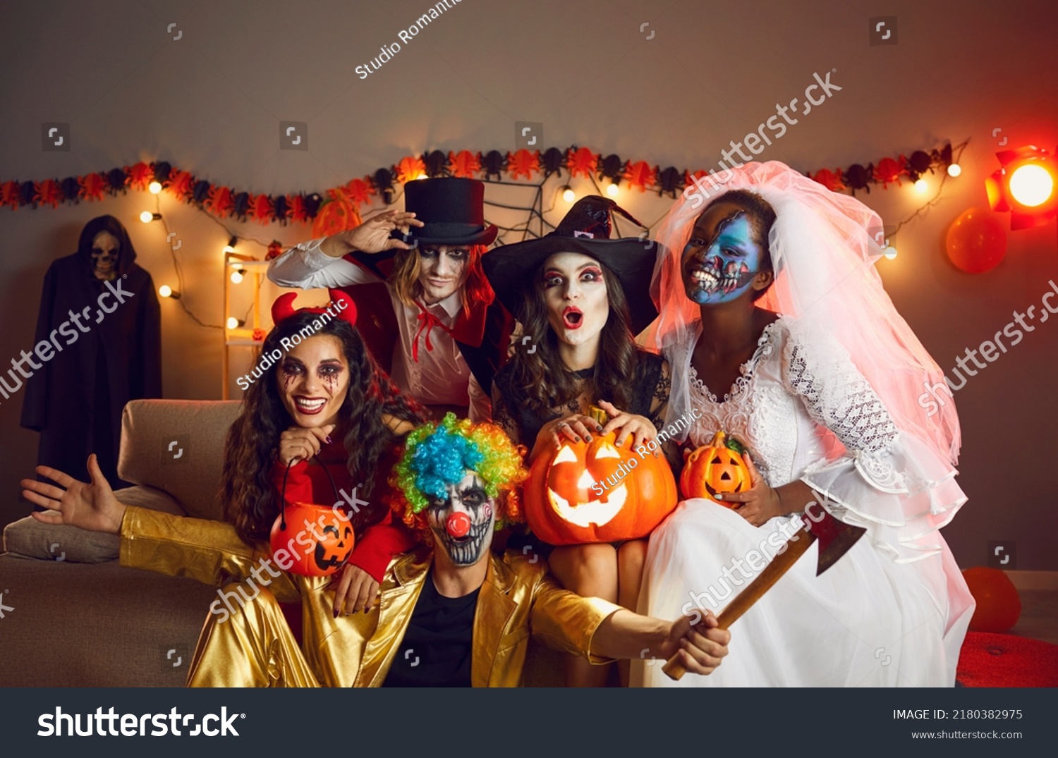 Happy adult friends in spooky costumes posing for funny group photo. Cheerful young people dressed as crazy witch, devil, vampire, dead bride and clown with axe having fun together at Halloween party #2180382975