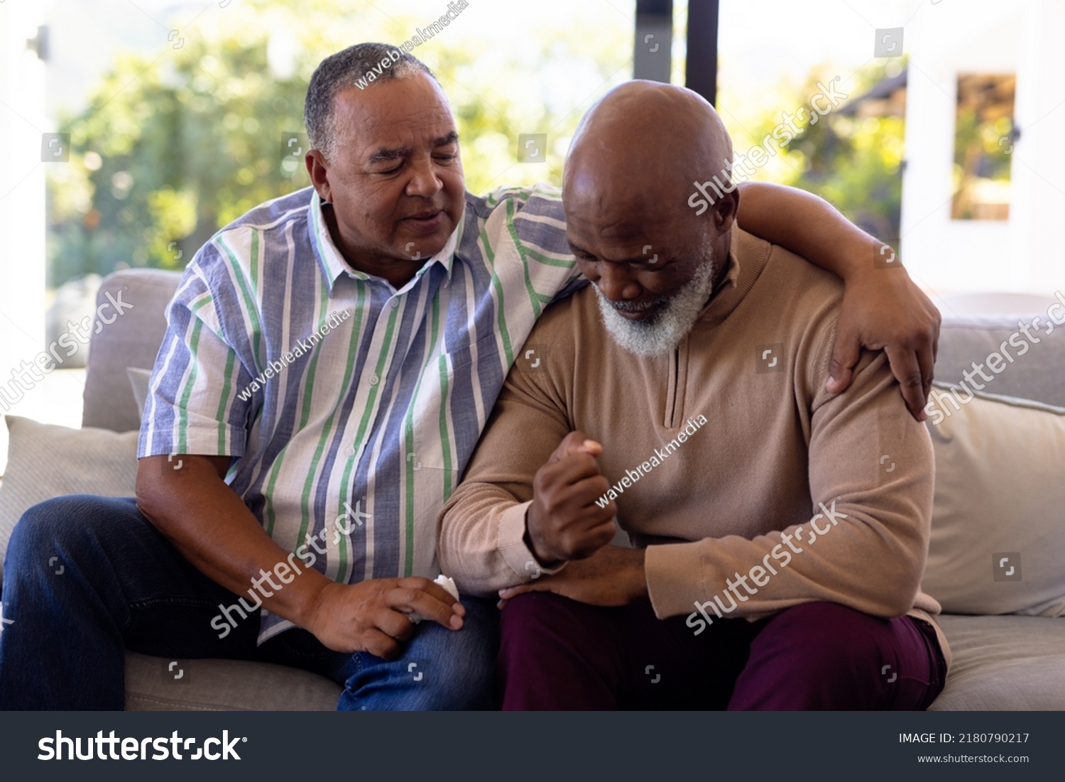 Multiracial senior man consoling male friend crying while sitting on sofa in nursing home. Sadness, togetherness, comfort, unaltered, emotional stress, support, assisted living, retirement concept. #2180790217