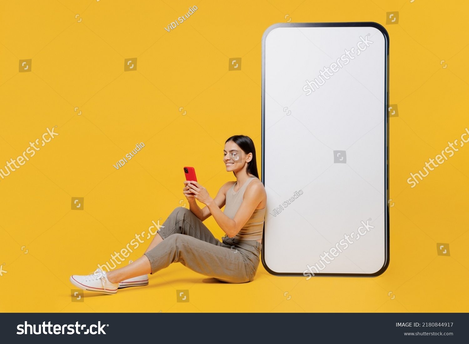 Full size smiling happy young latin woman she wear tank shirt sit near big huge blank screen mobile cell phone with workspace copy space mockup area use smartphone isolated on plain yellow backround #2180844917
