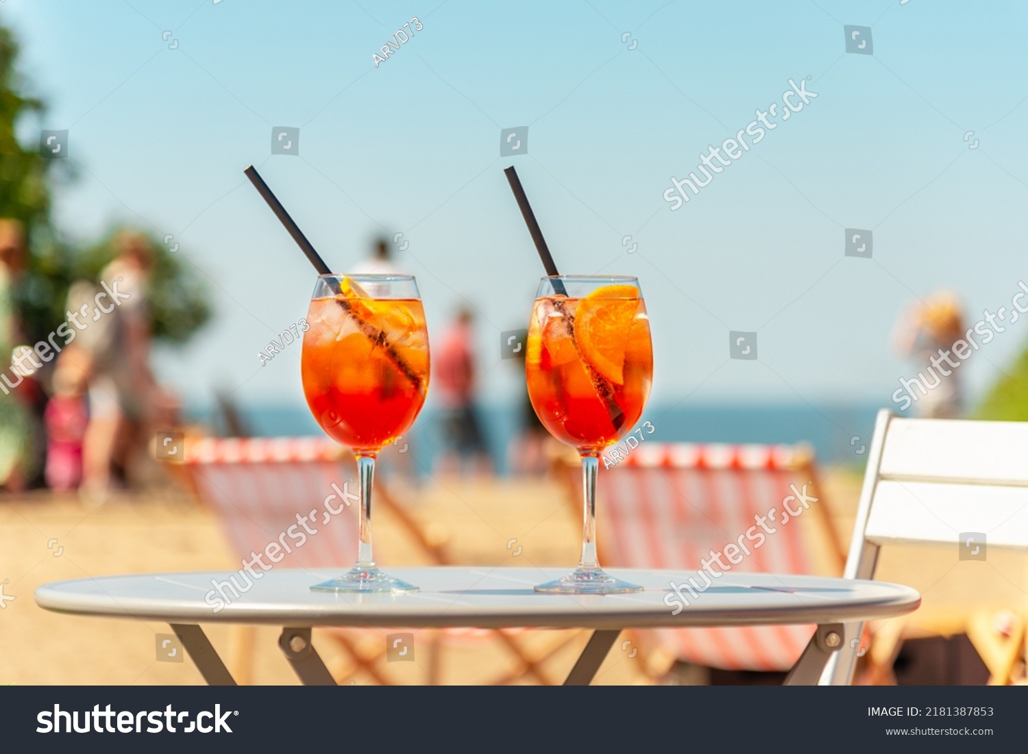 Two glasses of orange spritz aperol drink cocktail on table outdoors with sea and trees view blurred people background. #2181387853