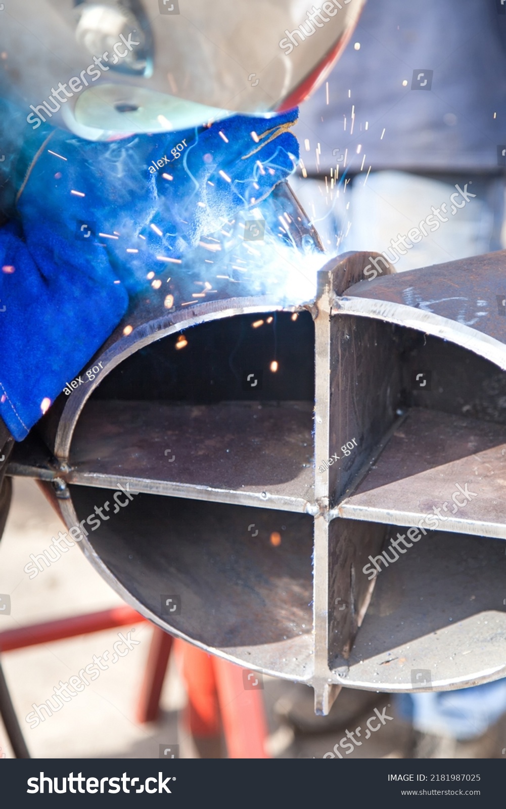 A professional welder in a protective suit and mask produces the connection of pipes with the help of a welding machine on the construction site. #2181987025