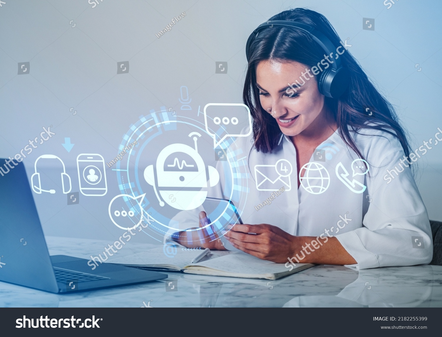 Businesswoman in headphones smiling, using phone, digital chat bot hud hologram. Office table with laptop and notebook. Concept of helpdesk #2182255399