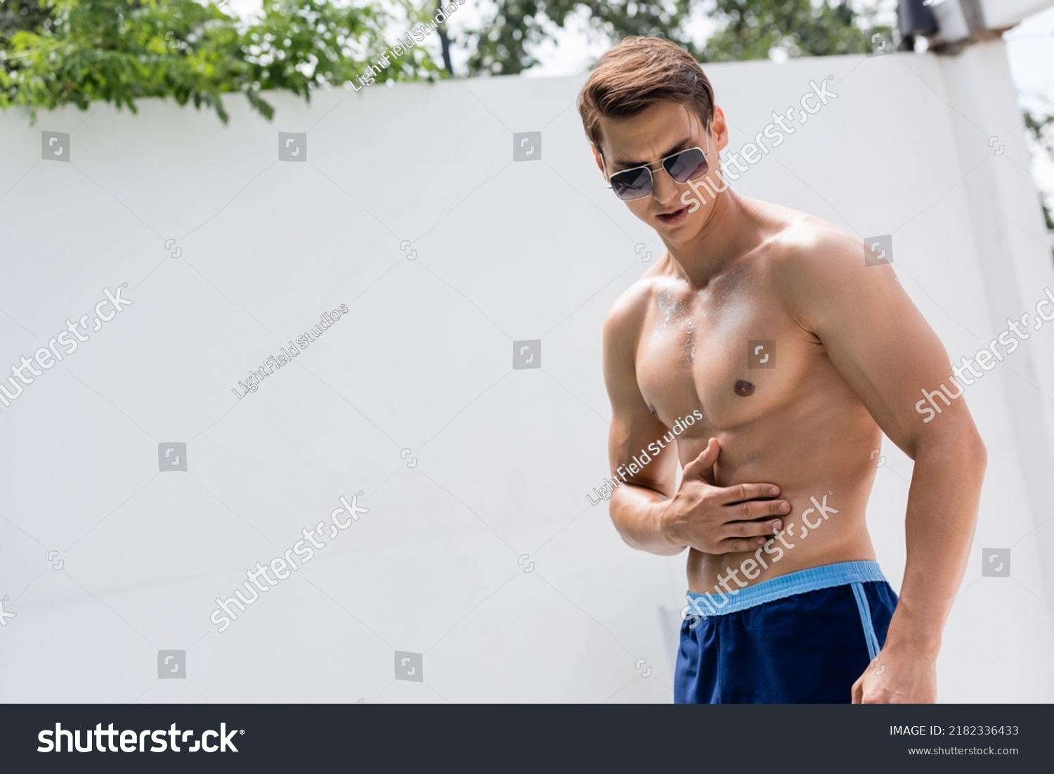 man in sunglasses and swimming trunks applying sunscreen on muscular torso #2182336433