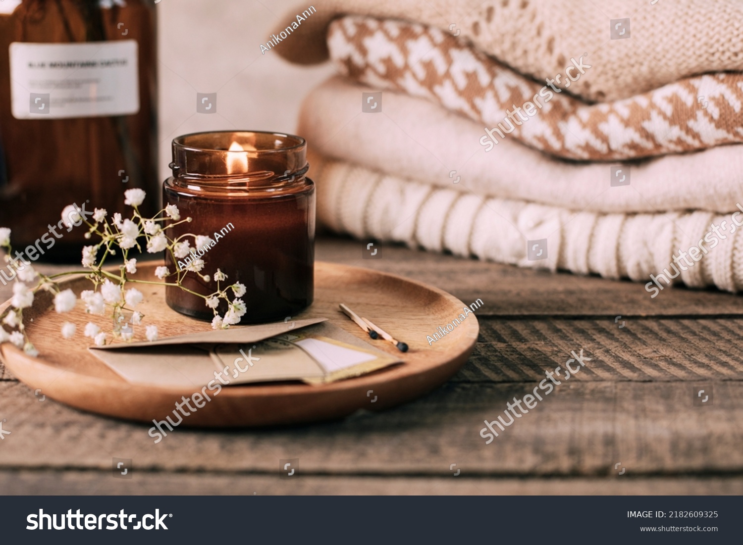 Burning candle in small amber glass jar, flowers of gypsophila and stack knitted seasin sweaters. Cozy lifestyle, hygge concept #2182609325