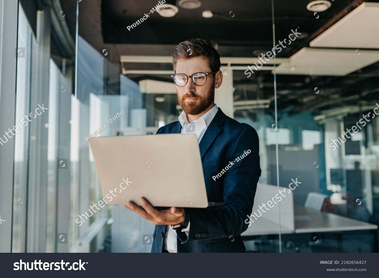 Business person. Successful middle aged businessman with laptop in hands standing in modern office interior. Serious male entrepreneur in formal suit working on computer #2182656427