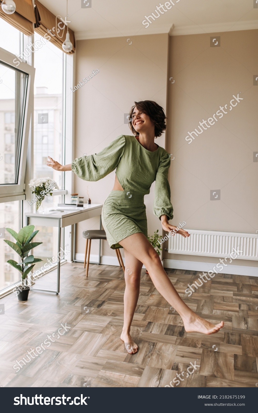 Full-length view of cheerful pretty woman staying in bright room . Caucasian short hair wear green dress smiling and looking at window. Lifestyle, leisure life concept  #2182675199