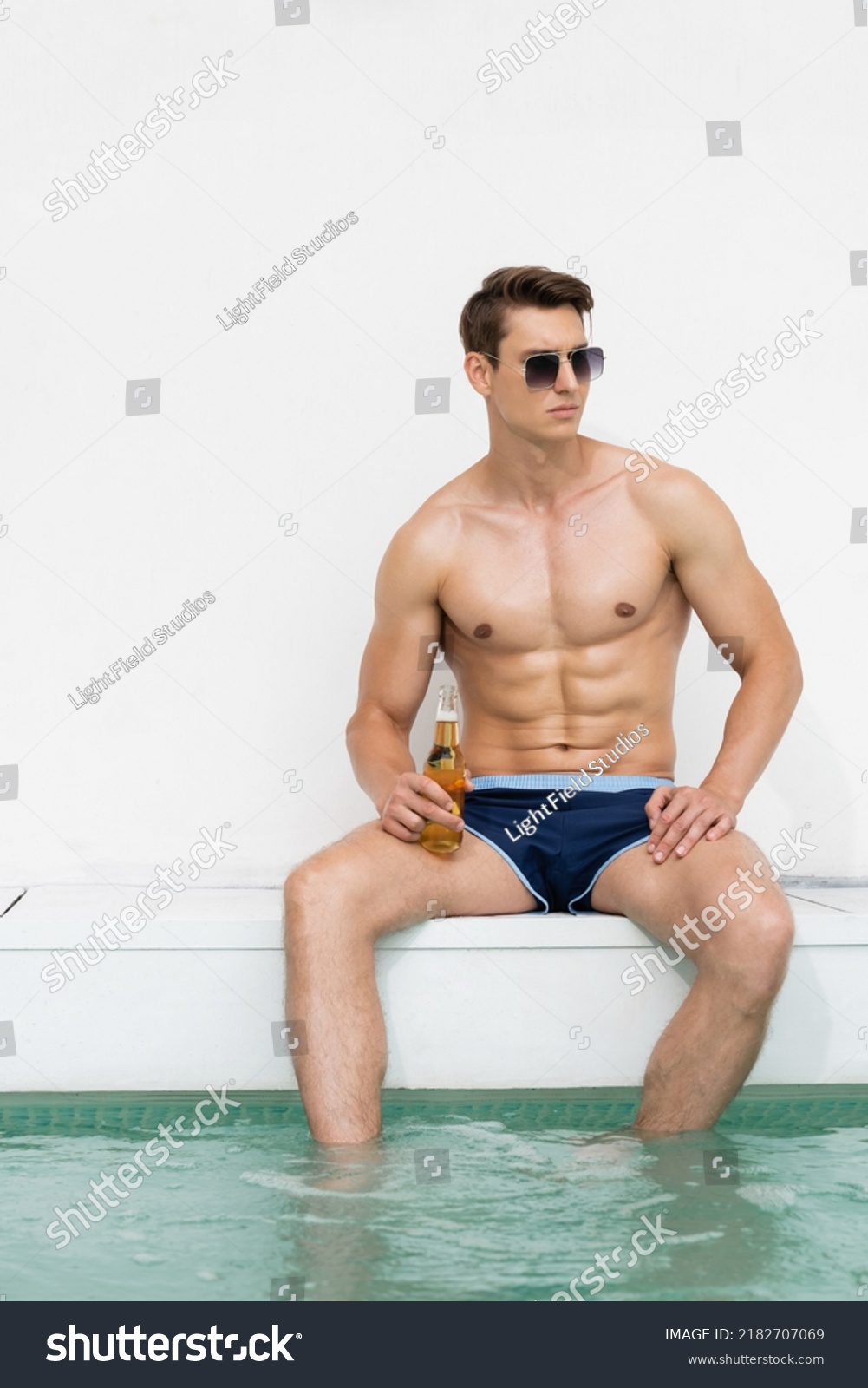 muscular man in swimming trunks and sunglasses sitting at poolside with bottle of beer #2182707069