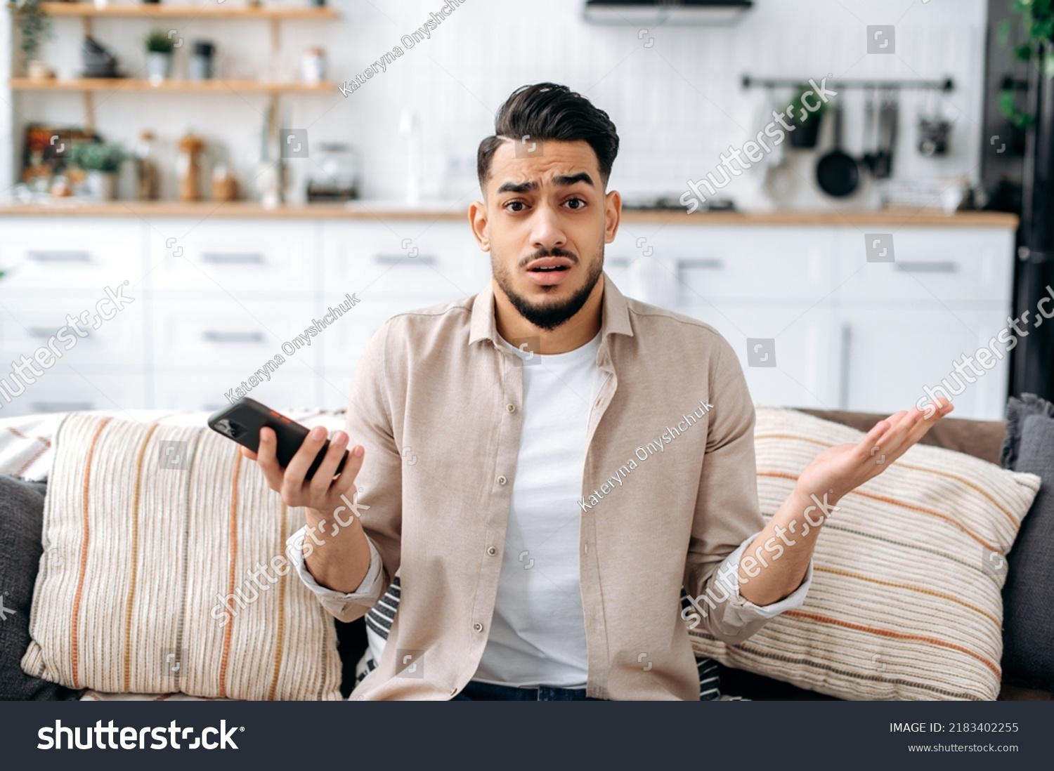 Confused puzzled indian or arabian guy in casual clothes, sits on a sofa in an interior living room, holds a smartphone in his hand, looks questioningly at the camera, spreading his arms around #2183402255