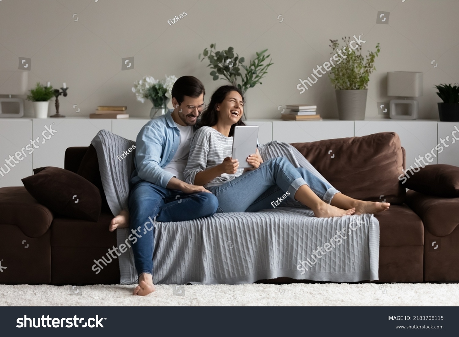 Happy 30s couple relaxing on soft comfortable couch, enjoying leisure time and entertainment in cozy home interior, using tablet, reading book, watching movie, laughing and hugging. Wide shot #2183708115