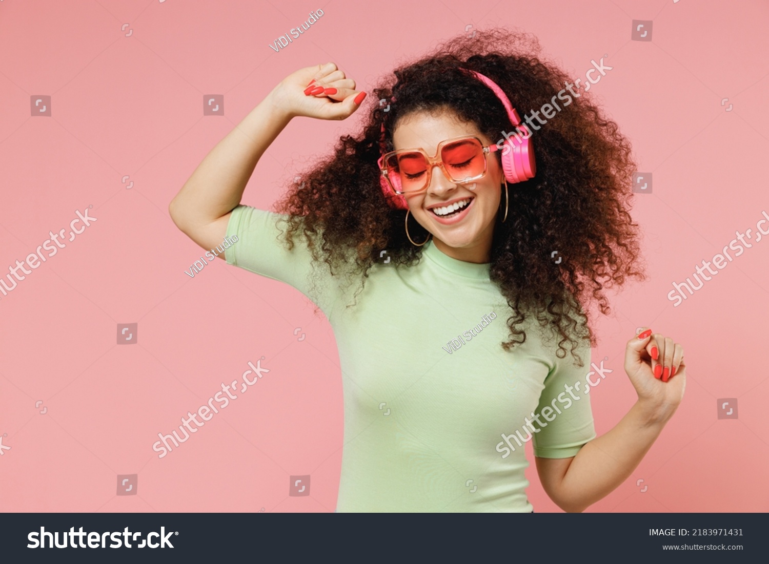 Bright fun young curly latin woman 20s wears mint t-shirt sunglasses listen music in headphones dance have fun rest relax fooling around isolated on plain pastel light pink background studio portrait #2183971431
