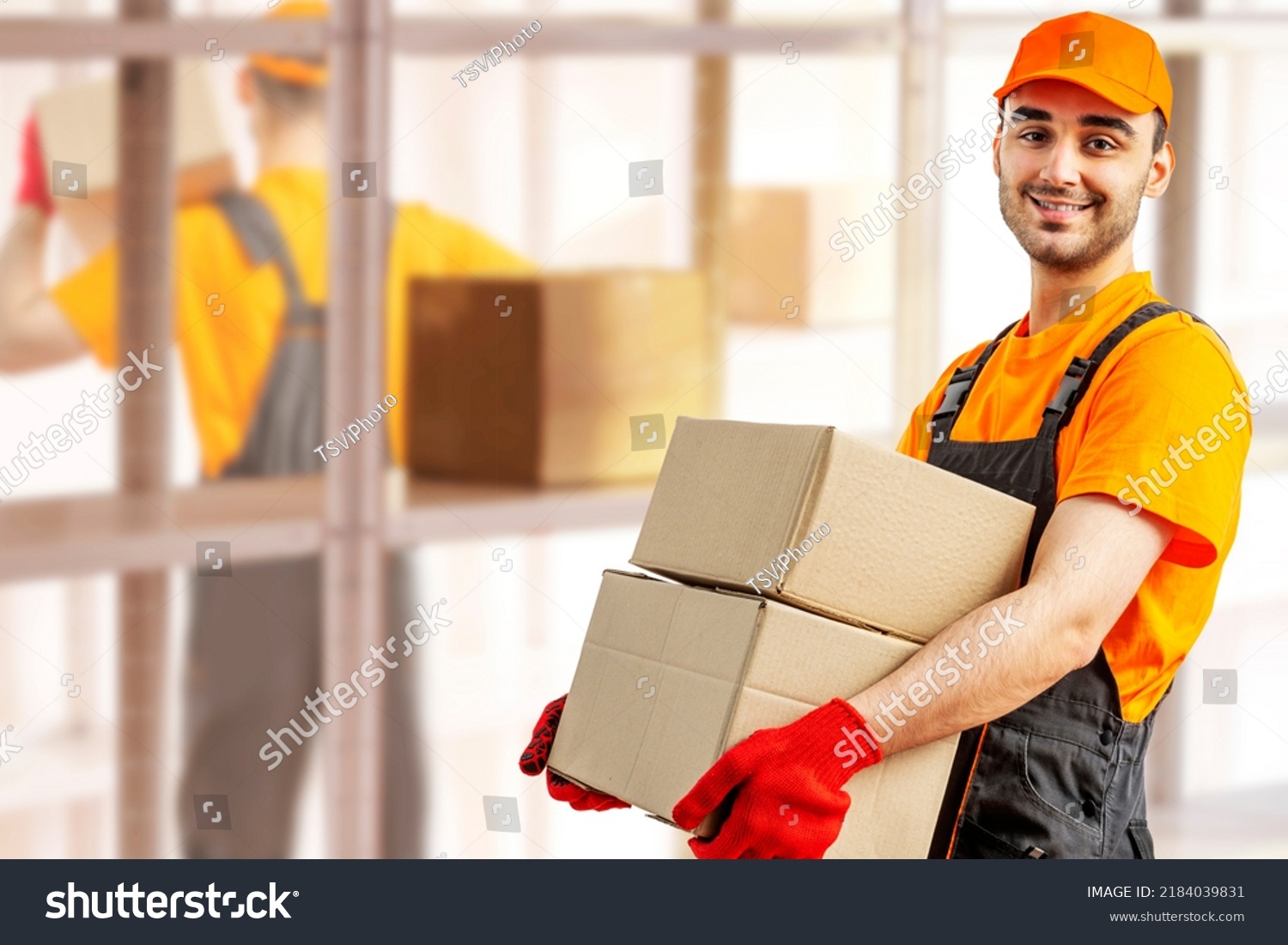 Young man holding cardboard package working in warehouse among racks and shelves. Delivery man with box. Staff laborer, orange uniform cap, t-shirt, coveralls service moving delivering orders goods.  #2184039831