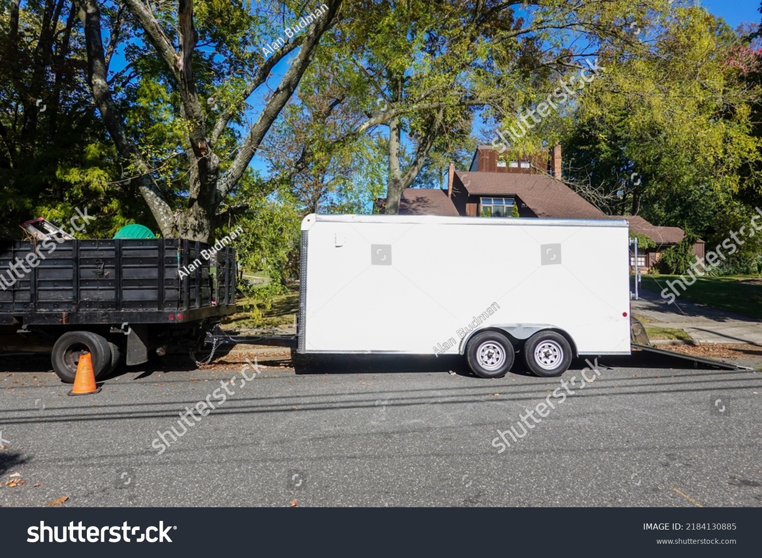 A landscaping truck with a long white enclosed trailer trailer seen on a shady residential asphalt street #2184130885