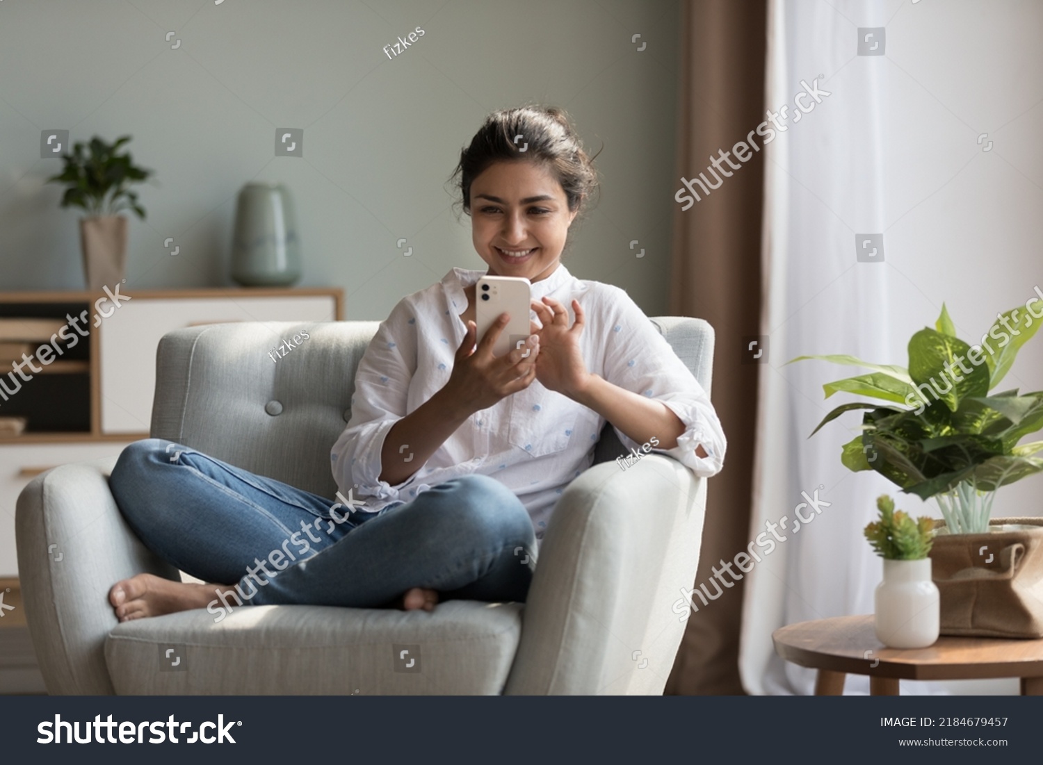 Happy pretty millennial Indian girl relaxing at home, resting in armchair, typing on smartphone, using online app, software, shopping on Internet, making video call. Mobile phone communication #2184679457
