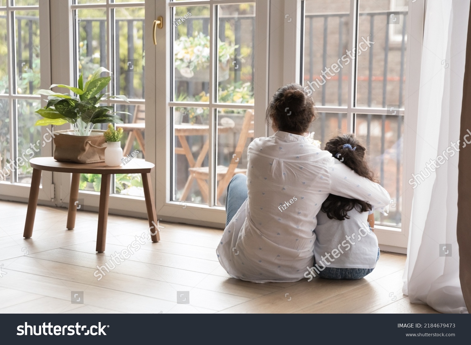 Back view of black haired young mom and little daughter girl sitting on floor at home, hugging with love, care, looking at panoramic window, cozy balcony. Motherhood, real estate concept #2184679473