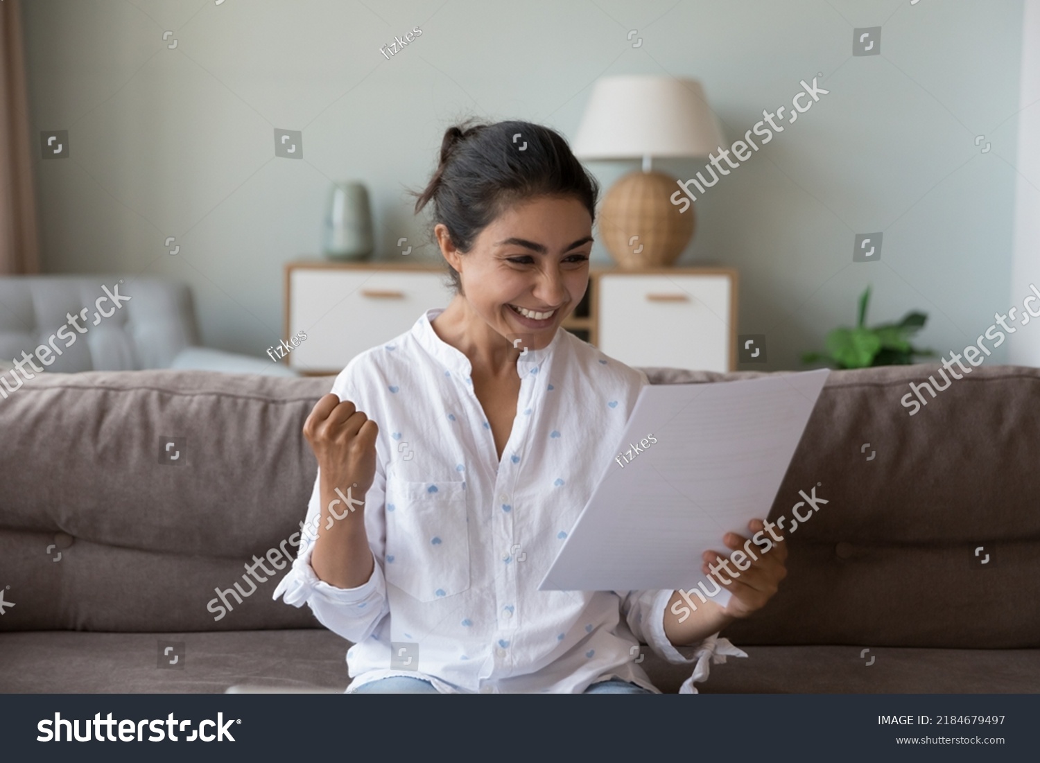 Excited cheerful young Indian woman getting good news letter, reading paper document at home, making winner yes hand, feeling happy, smiling, laughing, celebrating, success, achieve #2184679497