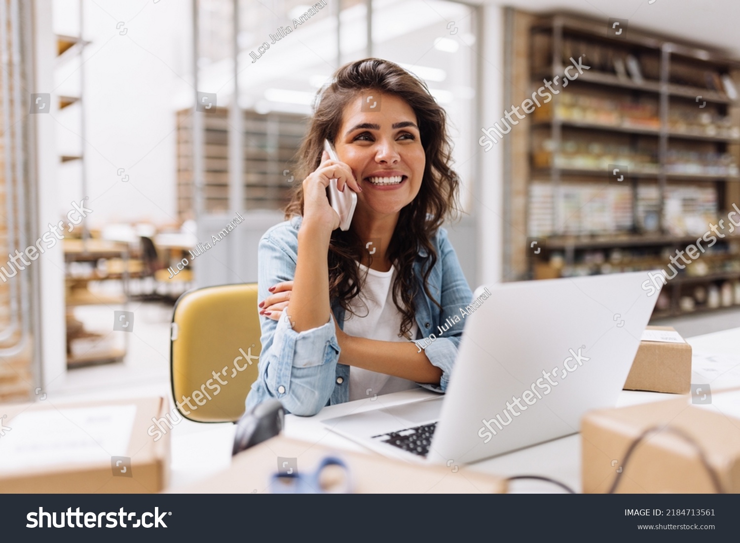Cheerful businesswoman speaking on the phone while working in a warehouse. Happy online store owner making plans for product shipping. Female entrepreneur running an e-commerce small business. #2184713561