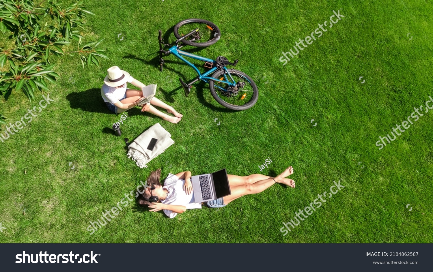 Young girls with bicycle in park, using laptop computer and headphones, two student girls studying online outdoors sitting on grass near bike, aerial drone view from above #2184862587