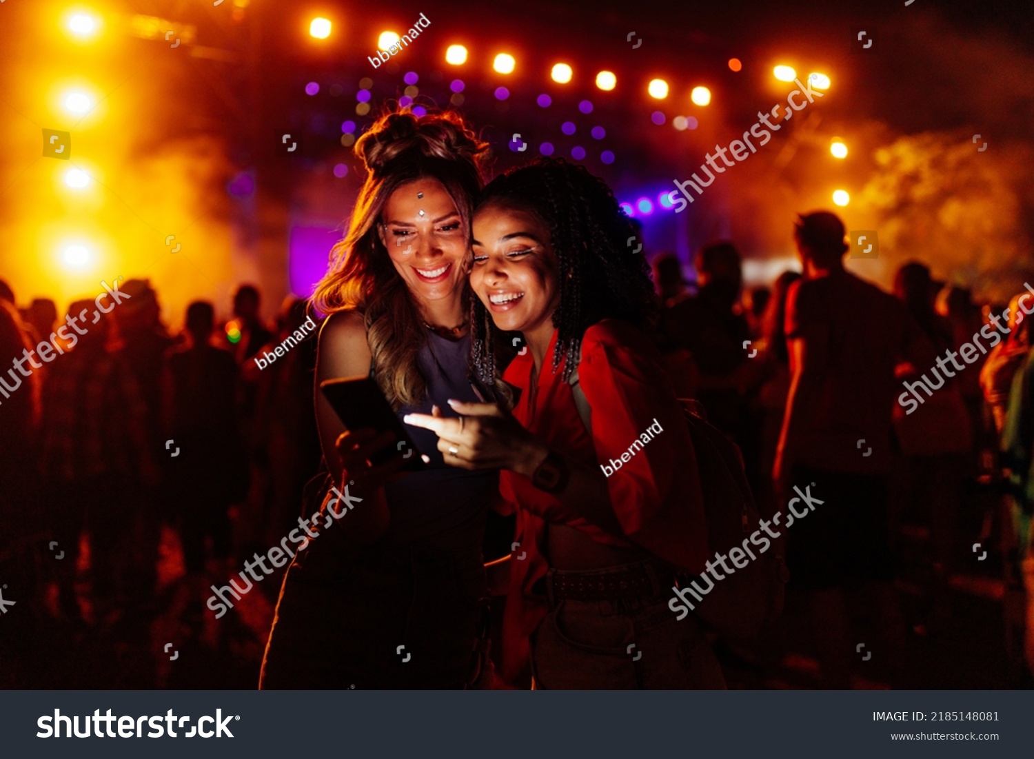 Two biracial women in a crowd at a concert using cellphone together #2185148081