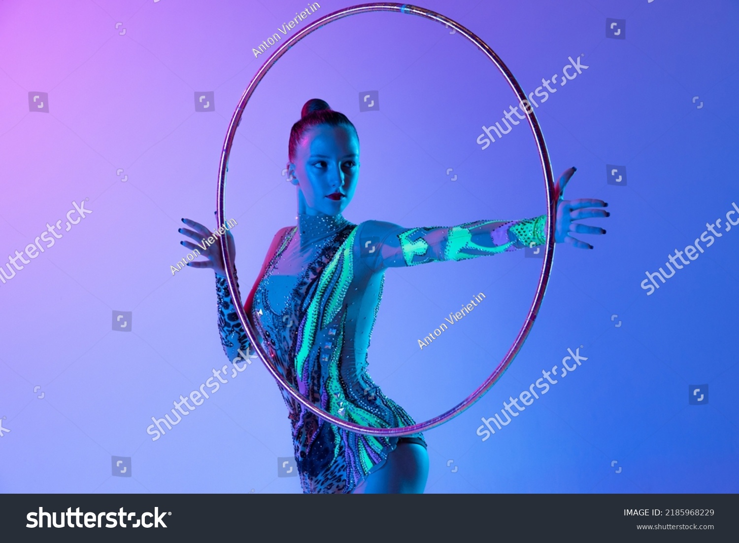 Portrait of young, muscular girl, female rhytmic gymnast training with hoop isolated over blue pink studio background in neon light. Concept of action, motion, sport life, motivation, competition. #2185968229