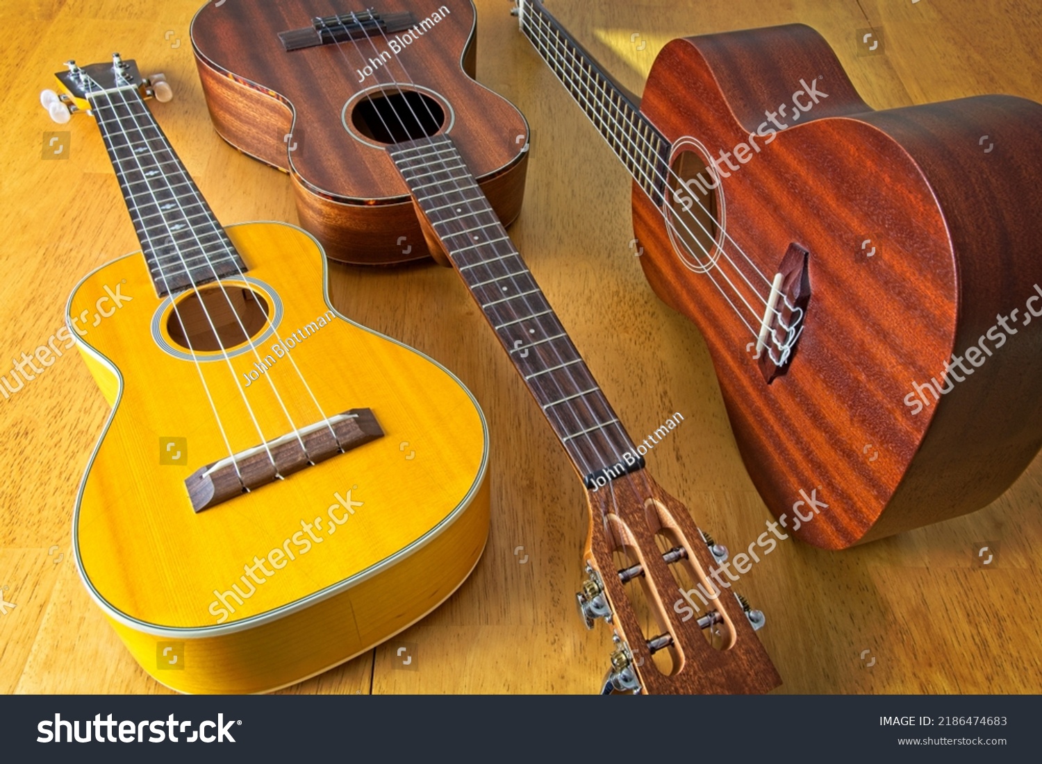 Collection of three ukuleles aligned on sun drenched wooden table top. High angle view. Side lit, showing texture of wood grains. #2186474683