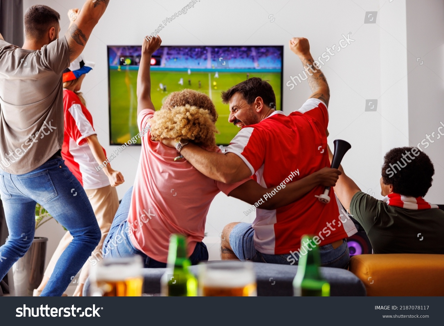 Group of young friends having fun watching football match on TV, drinking beer and cheering; football fans watching game at home celebrating after their team scoring a goal #2187078117