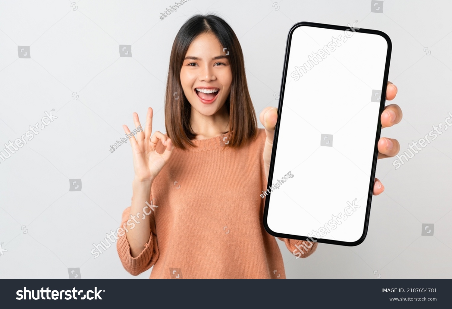 Beautiful Asian woman holding smartphone mockup of blank screen and shows ok sign on grey background #2187654781