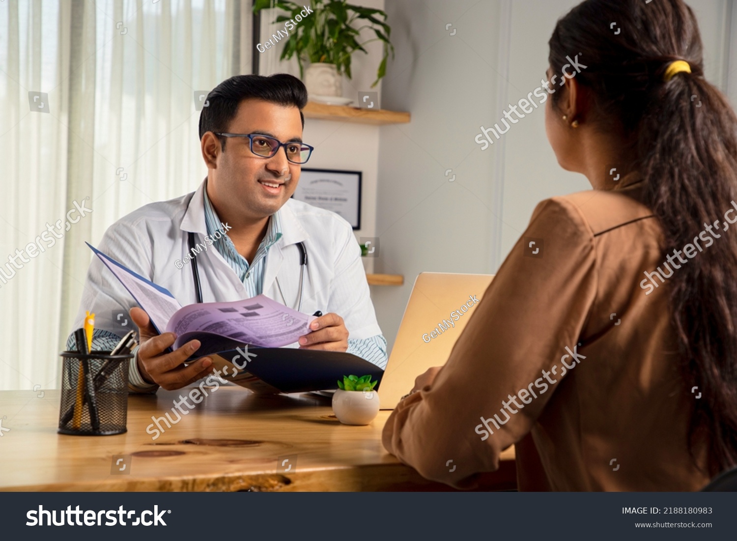 Asian Indian male physician or doctor wearing white apron and stethoscope holding a file of medical report is consulting or prescribing medicines to a Young Female patient in modern clinic or hospital #2188180983