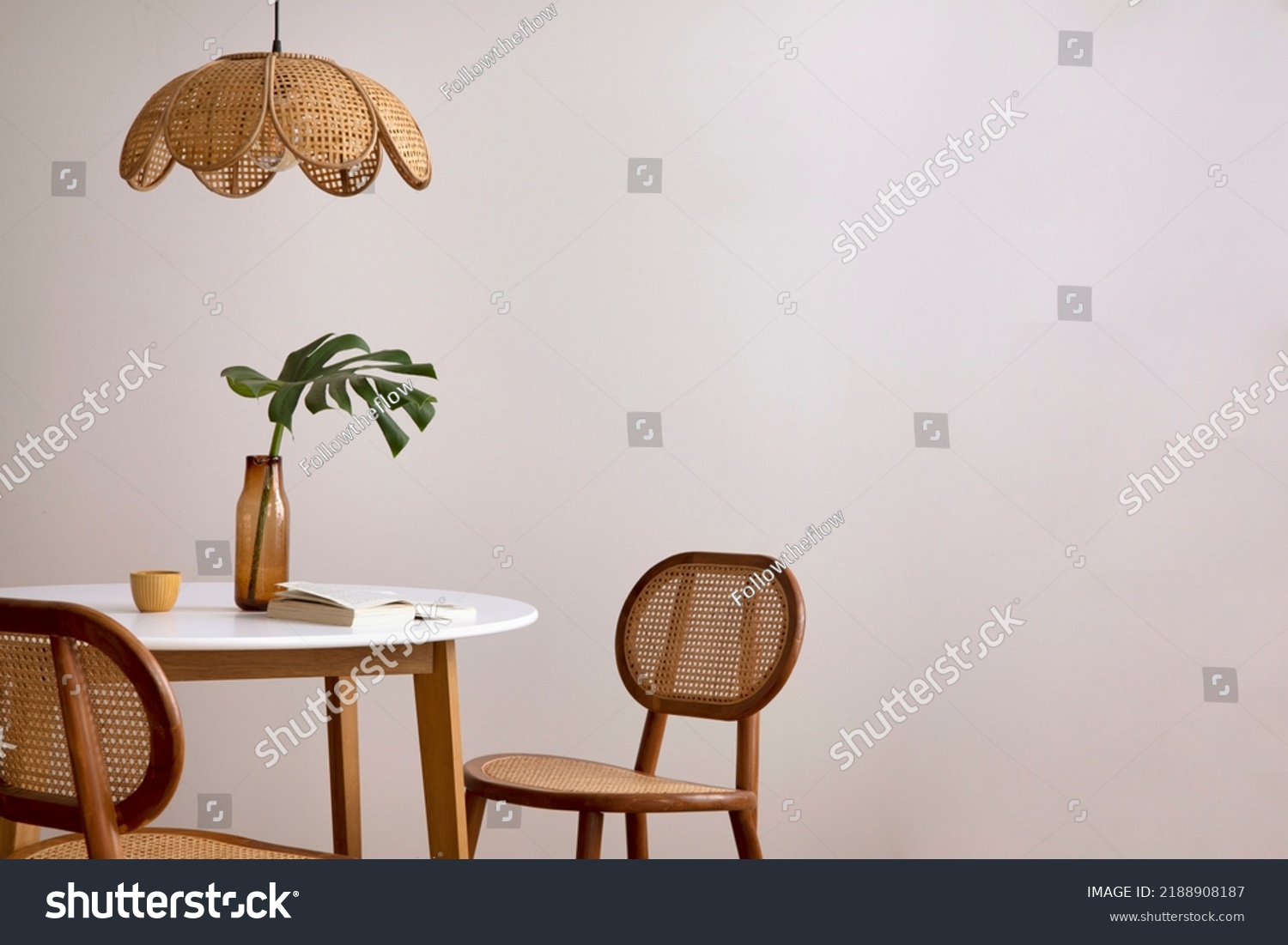 Minimalist composition of elegant kitchen space with round table, rattan chair, vase with leaf, pedant lamp and personal accessories. Minimalist home decor. Beige wall. Template.  #2188908187