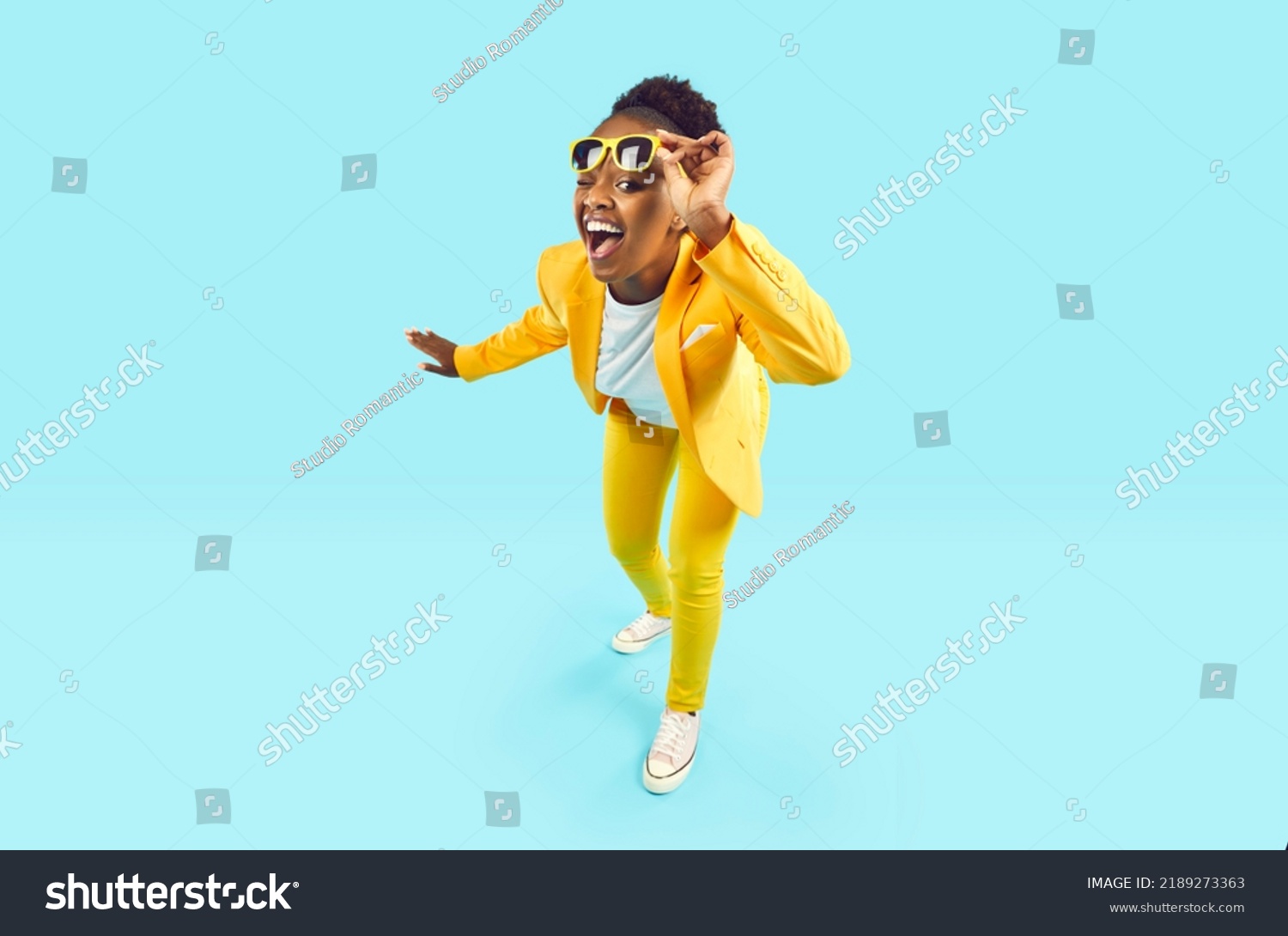 Full body happy cheerful Afro American woman wearing stylish yellow suit and trendy glasses standing isolated on blue background, looking at camera, winking her eye and smiling. Party, fashion #2189273363