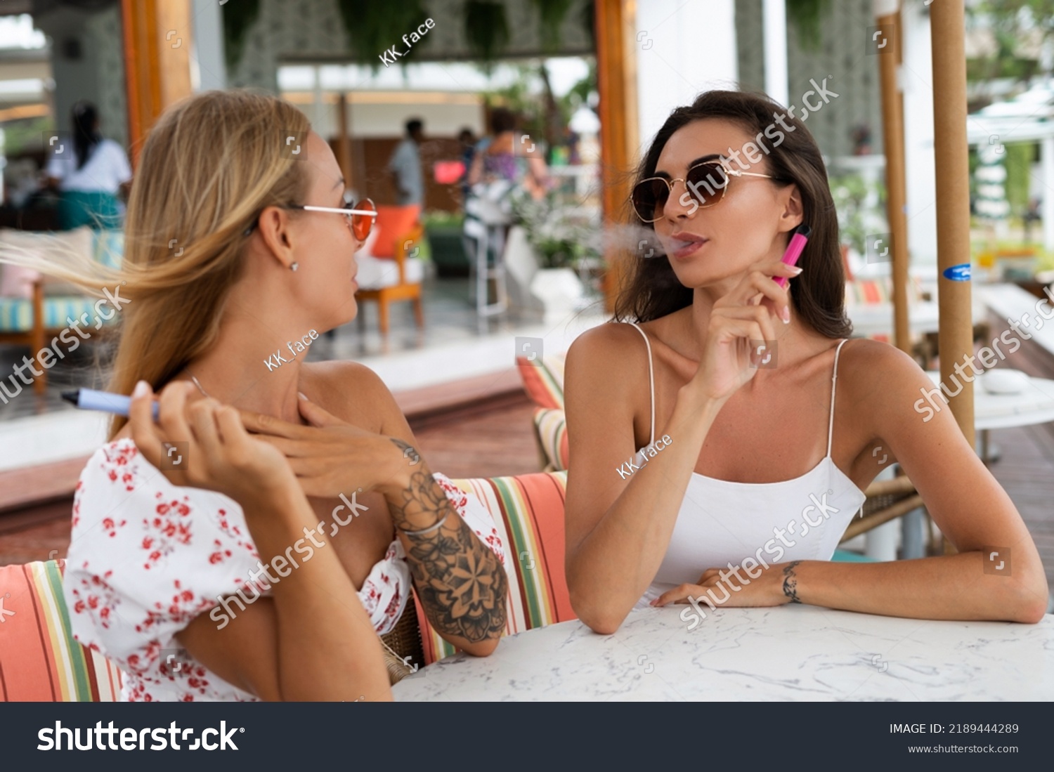 Two young beautiful smiling hipster female in trendy summer sundress.Smoking vapes electronic devices at outdoor cafe. Positive models having fun #2189444289