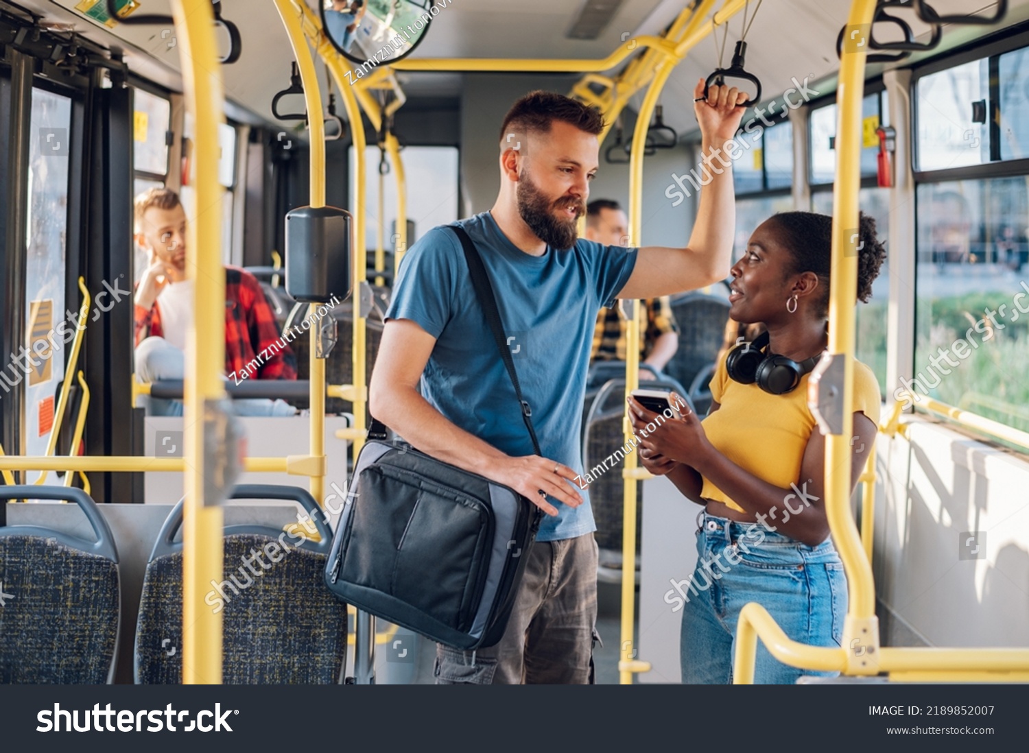 Cheerful diverse friends smiling and talking together and using a smartphone while riding a bus in the city and going to work. Standing in public transport, commuting by bus. Diverse romantic couple. #2189852007