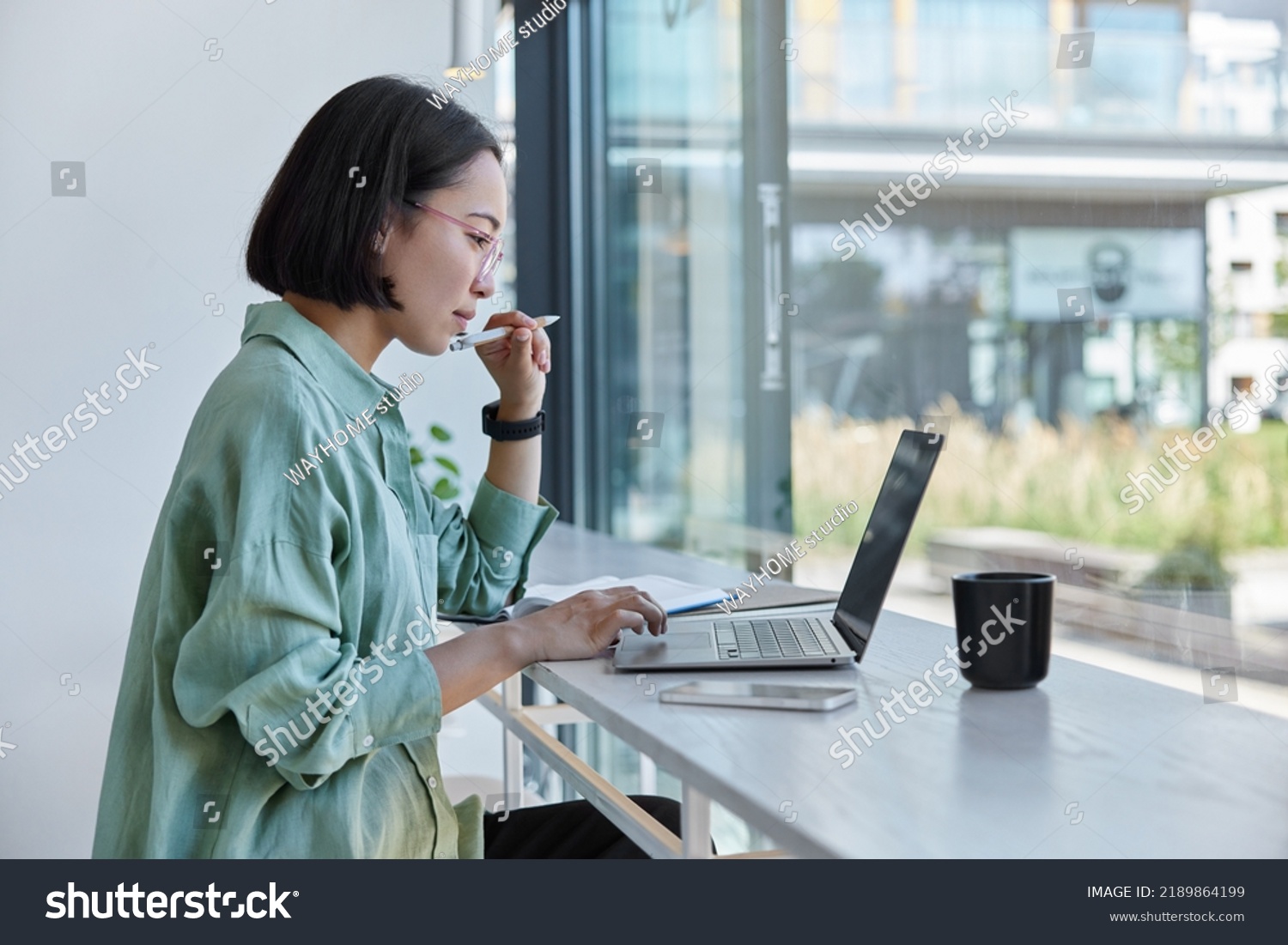 Millennial Asian student prepares for test or exam browses laptop studies in coffee house makes notes in notepad watches online webinar learns foreign language. E learning and online studying #2189864199