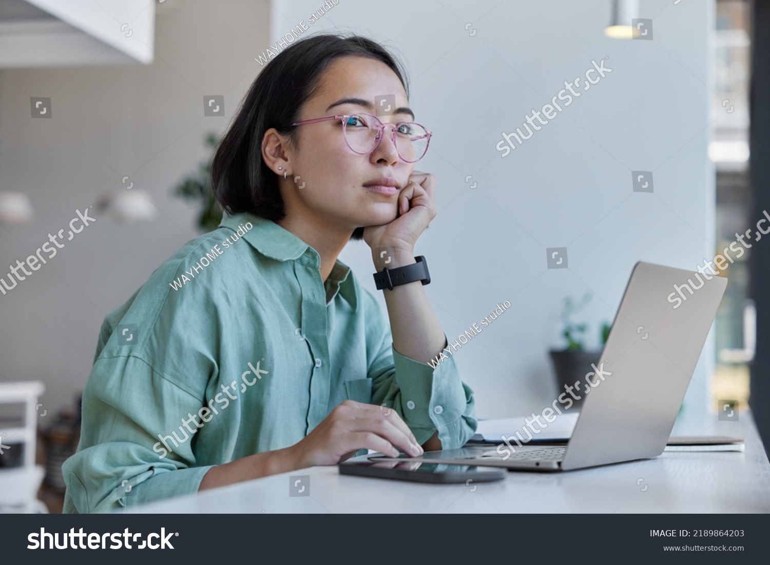 Thoughtful Asian woman works freelancer uses laptop computer smartphone has distance job poses against cozy interior indoors wears spectacles and shirt. Female digital nomad enjoys modern lifestyle #2189864203