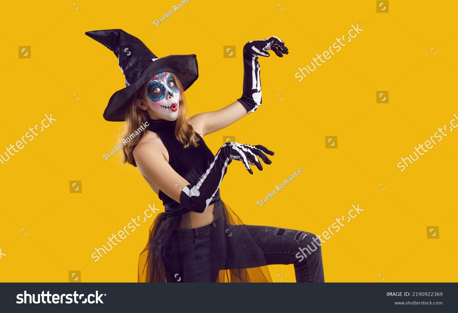 Portrait of funny child in Halloween disguise dancing isolated on yellow color background. Happy little girl in skeleton costume and witch hat, with skull makeup having fun at Halloween party #2190922369