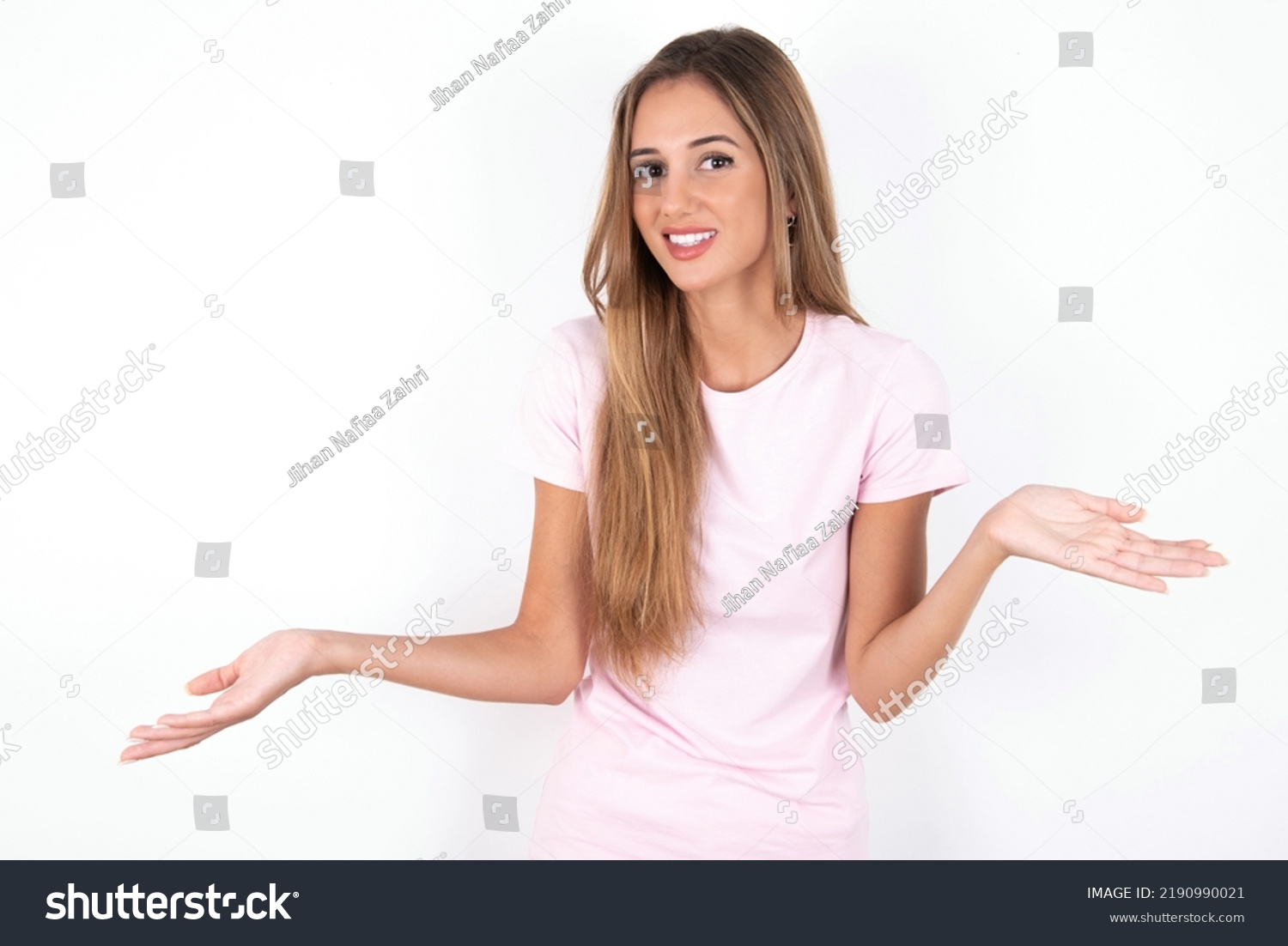 So what? Portrait of arrogant young beautiful woman wearing pink T-shirt over white background shrugging hands sideways smiling gasping indifferent, telling something obvious. #2190990021