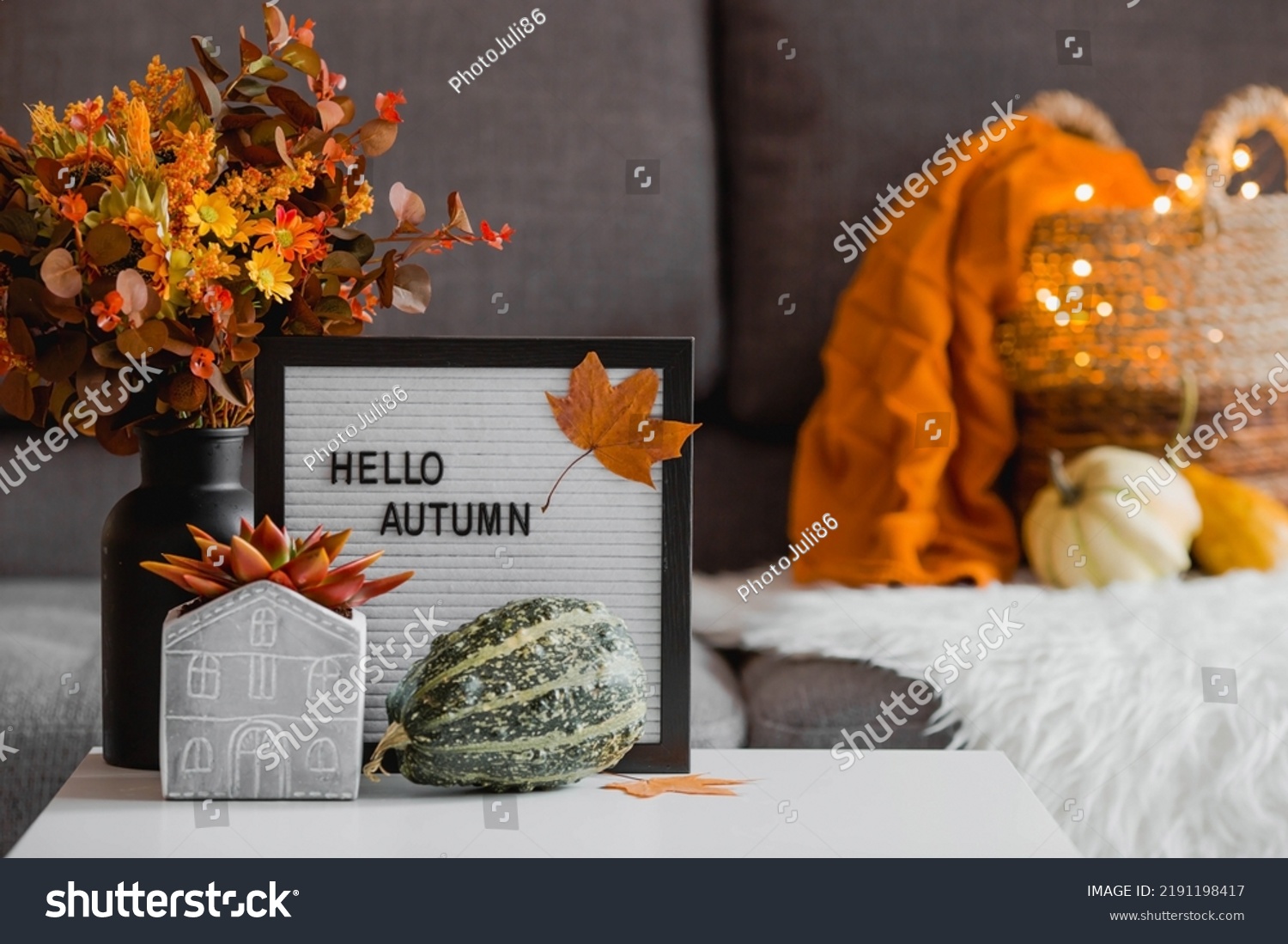 Cozy autumn concept. Home warmth in cold weather. Still-life. A blanket, pumpkins, flowers and the inscription home on the coffee table in the home interior of the living room. #2191198417