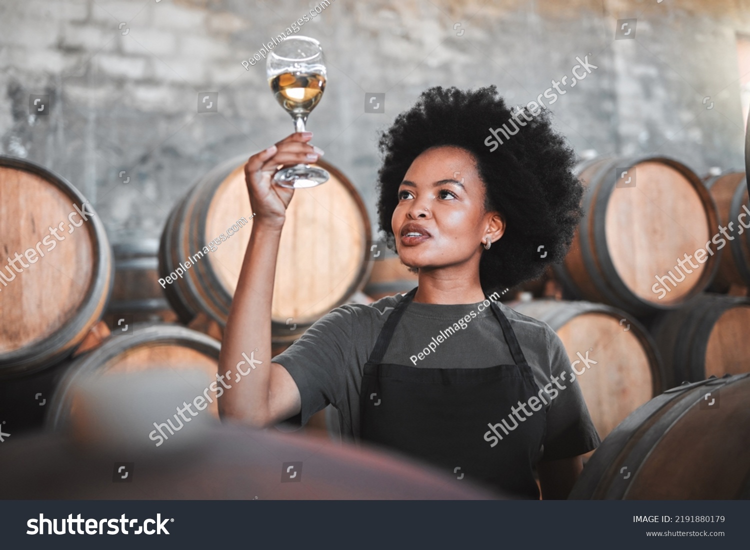 Black woman tasting wine at a winery, looking and checking the color and quality of the years produce. Young African American sommelier proud of the new addition, analyzing white wine in a cellar #2191880179