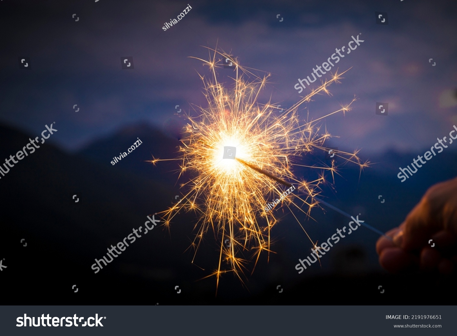 Hand holding a sparkler, or bengal fire stick, burning in outdoor setting, mountain landscape, at dusk. Holidays or magic background or wallpaper. #2191976651