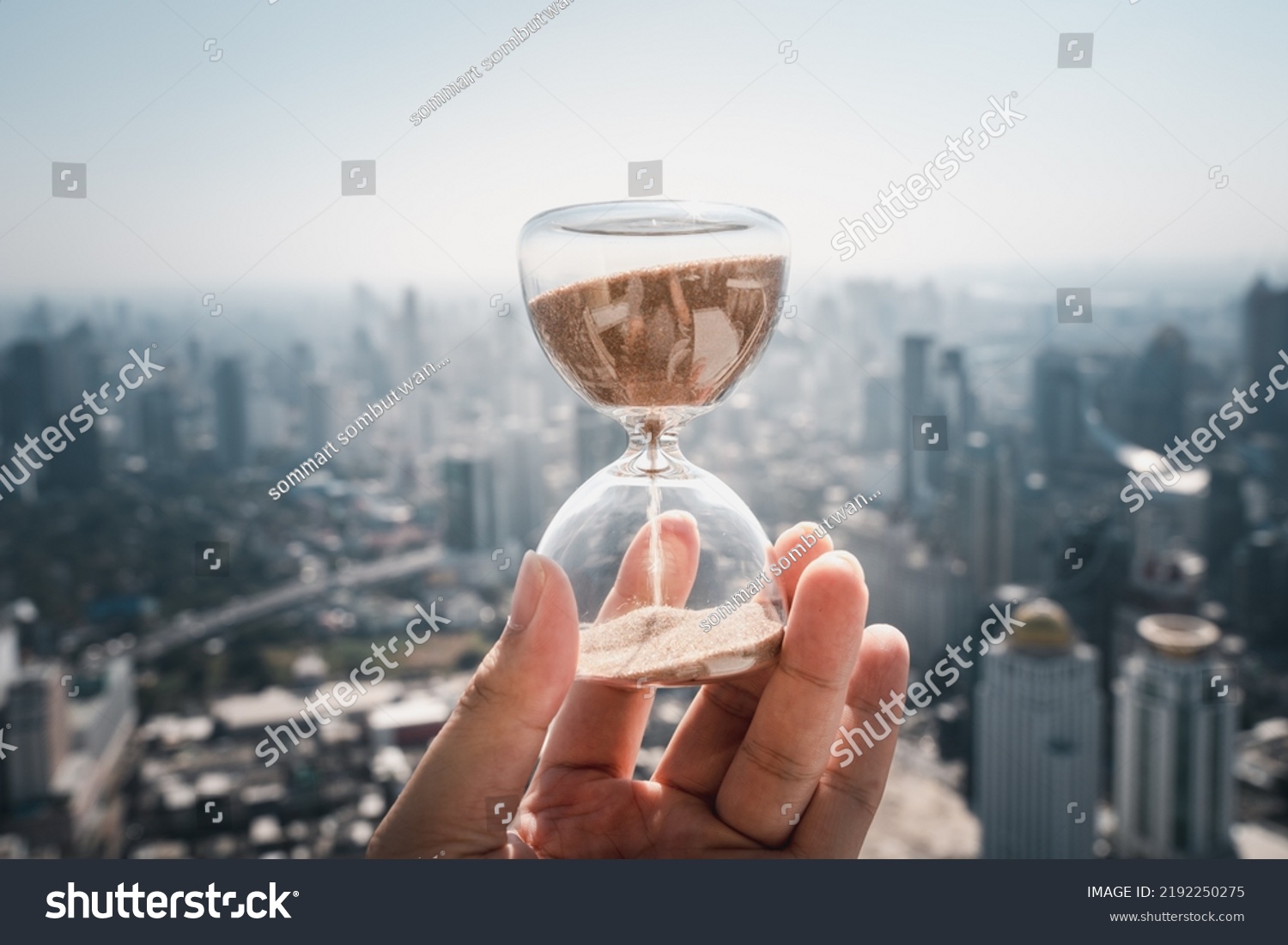 Hand holding a Hourglass with cityscape on panoramic skyline and buildings in the morning background with sun light. The concept of modern life, business, time, management and city life. #2192250275