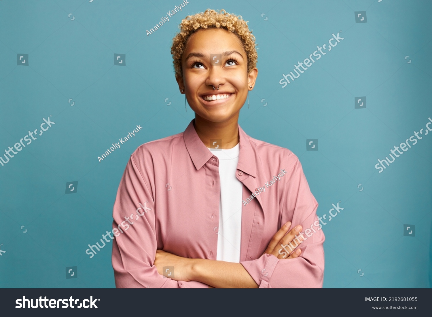 Portrait of pretty black girl of 20s looking up smiling biting lips, planning something, remembering funny moments, thinking about her boyfriend, standing with crossed hands on blue background #2192681055